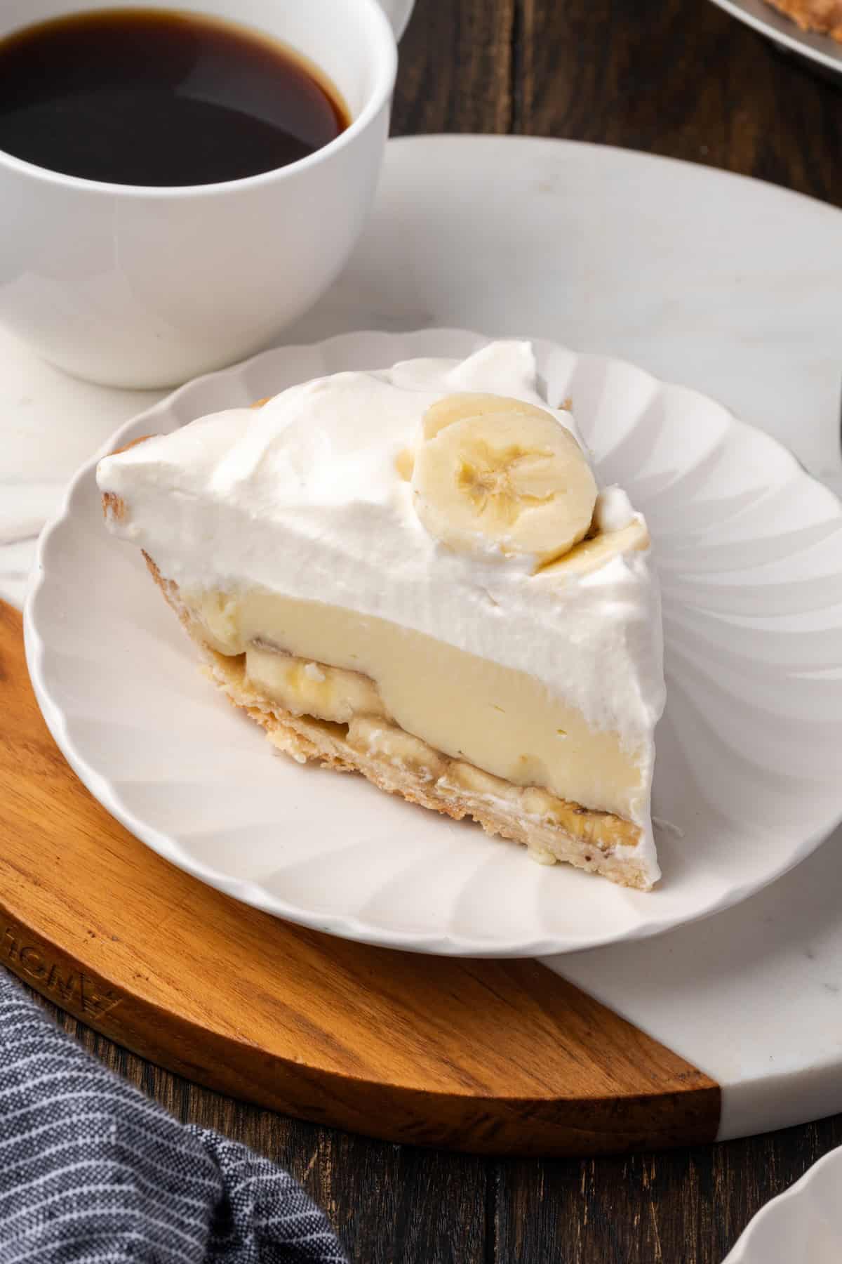 This banana cream pie recipe is a classic dessert with a rich vanilla custard layered with heaps of whipped cream and sweet banana slices. A slice of banana cream pie on a plate with a cup of coffee in the background.