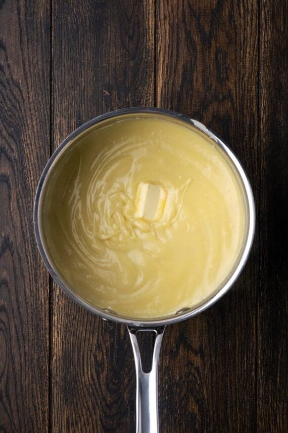 This banana cream pie recipe is a classic dessert with a rich vanilla custard layered with heaps of whipped cream and sweet banana slices. Butter added to custard in a saucepan.