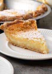 A slice of buttermilk pie dusted with powdered sugar on a plate with the rest of the pie in the background.