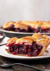 Side view of a slice of cherry pie on a white plate with the rest of the pie in the background.