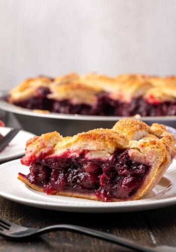 Side view of a slice of cherry pie on a white plate with the rest of the pie in the background.