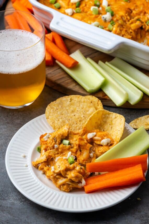 Chicken wing dip served on a plate next to chips and crudités, with a glass of beer and the rest of the dip in the background.