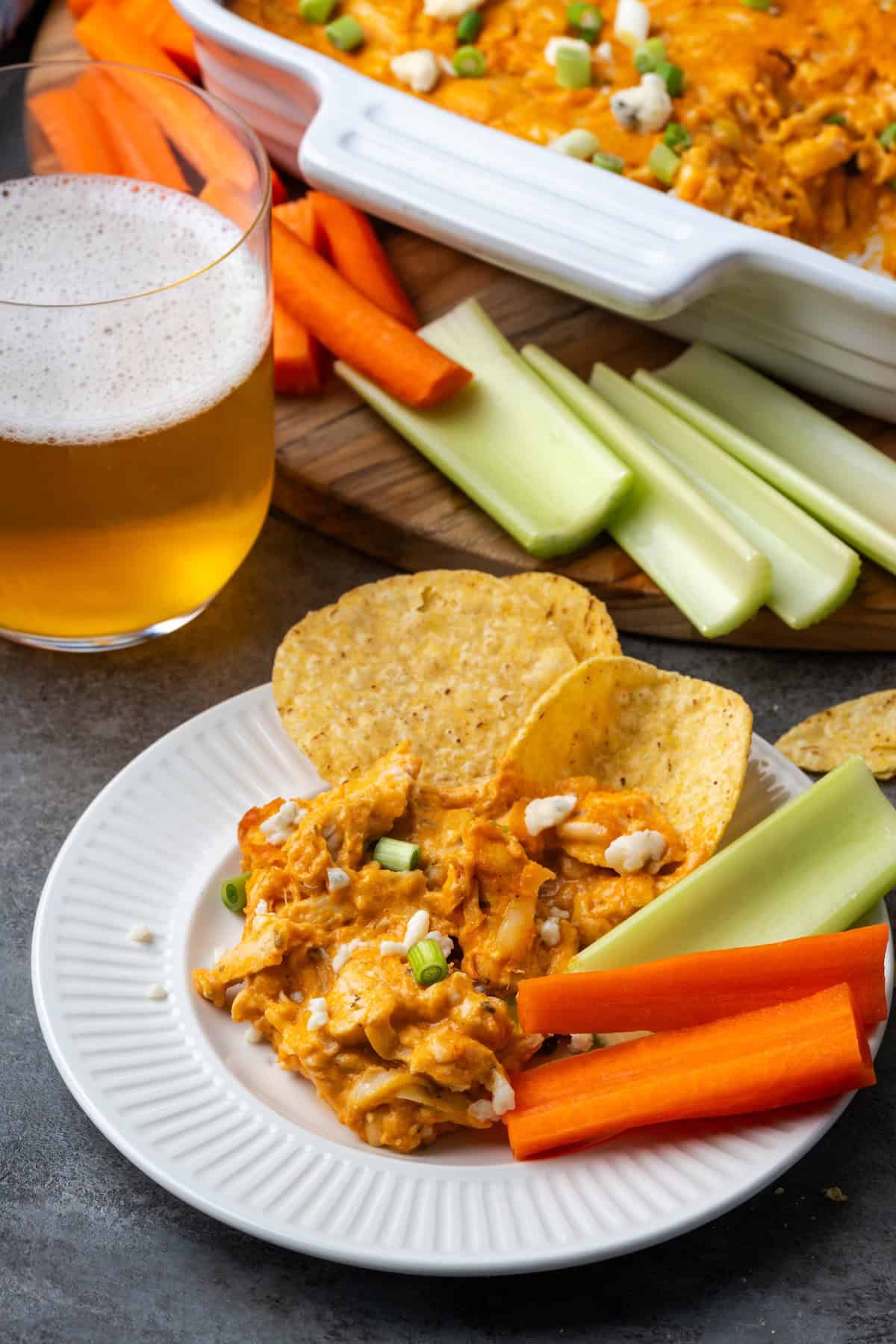 Chicken wing dip served on a plate next to chips and crudités, with a glass of beer and the rest of the dip in the background.