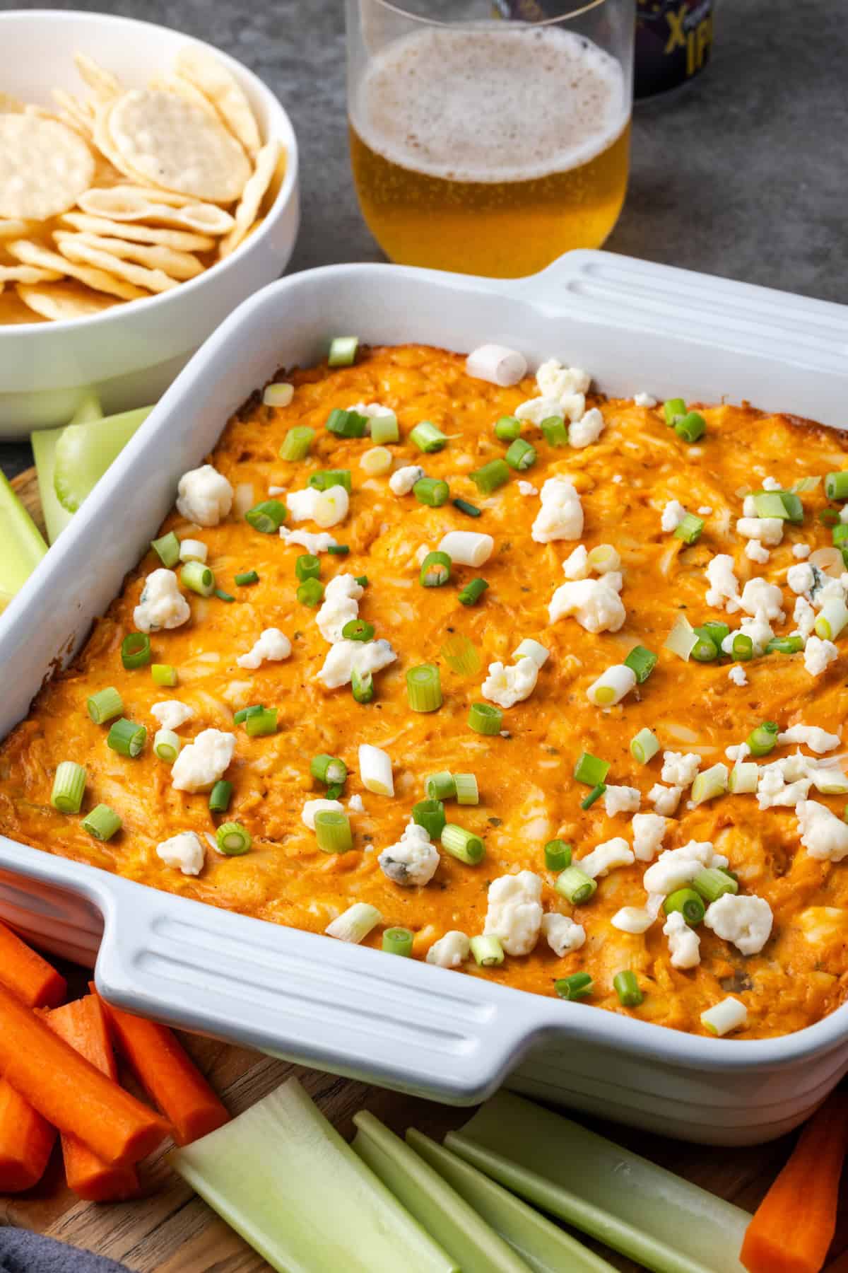 Chicken wing dip garnished with crumbled blue cheese and green onions in a baking dish, surrounded by carrot and celery sticks, with a bowl of chips and glass of beer in the background.