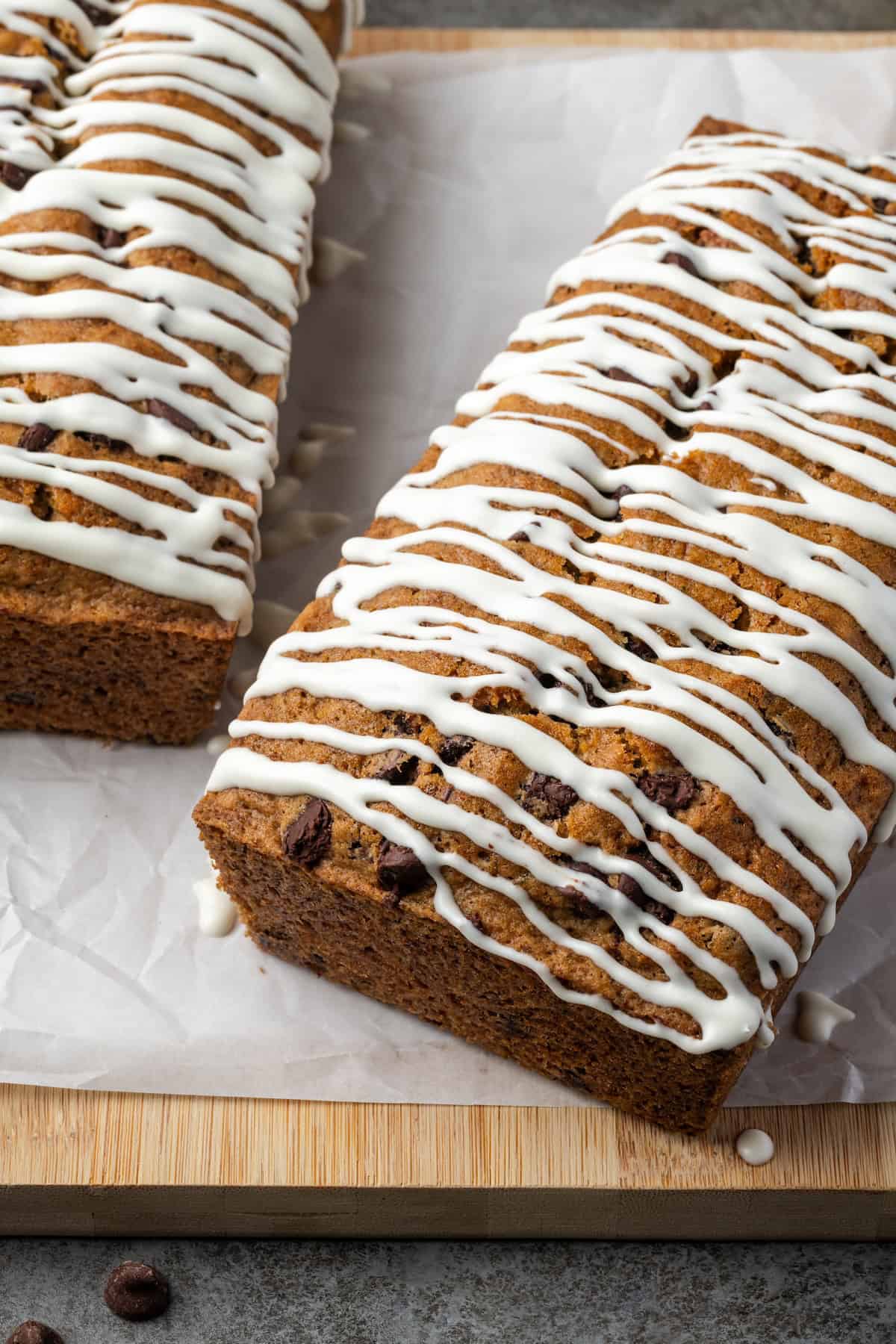 Two chocolate chip zucchini bread loaves drizzled with glaze on a wooden cutting board.