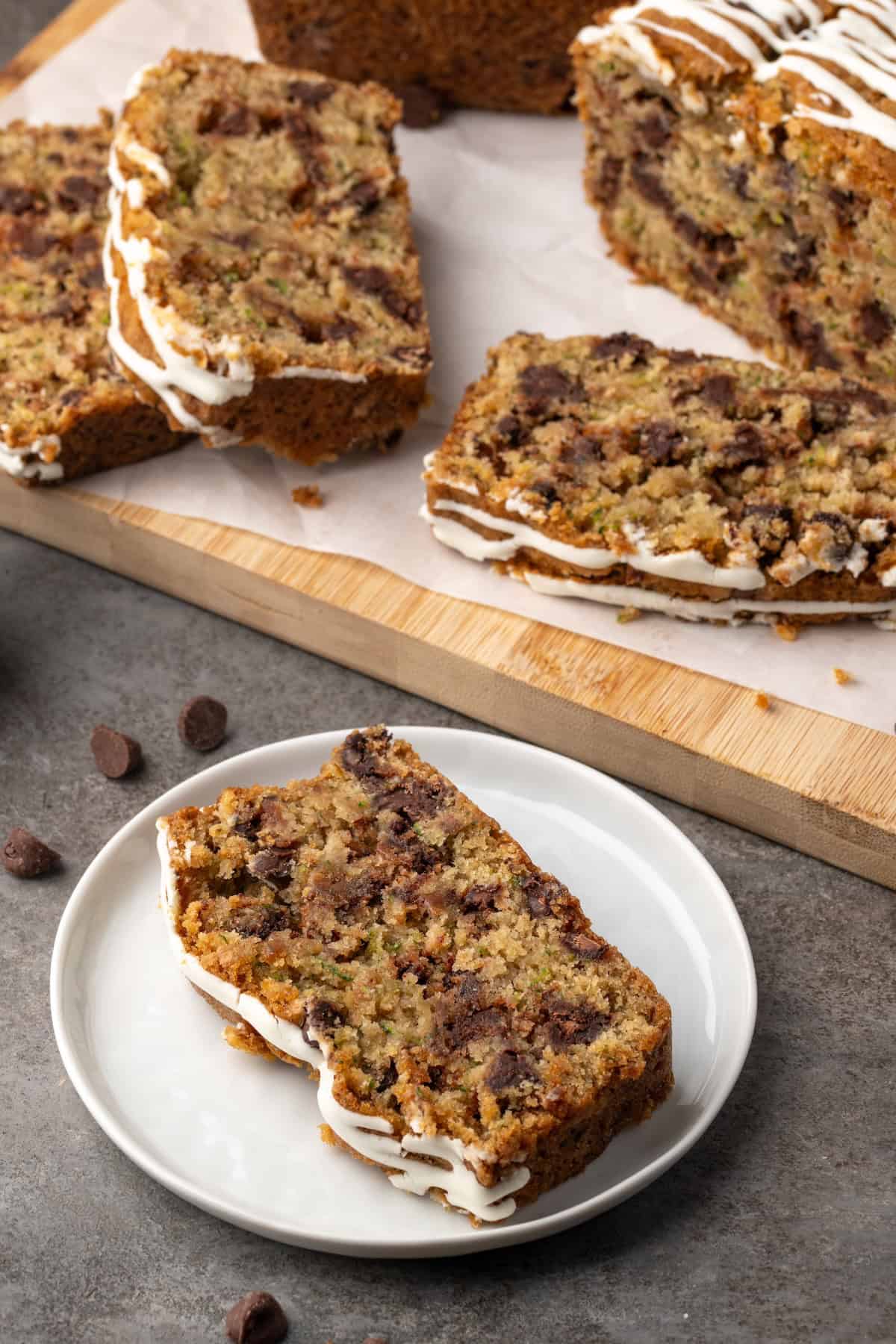 A slice of chocolate chip zucchini bread on a white plate with a loaf cut into slices on a wooden cutting board in the background.