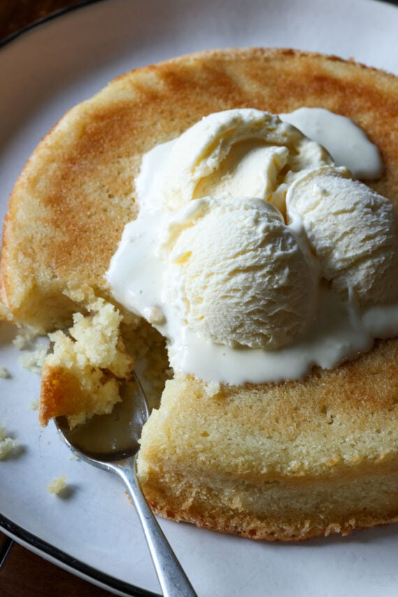 A 9 inch butter cake topped with vanilla ice cream on a white plate with bites taken out with a spoon