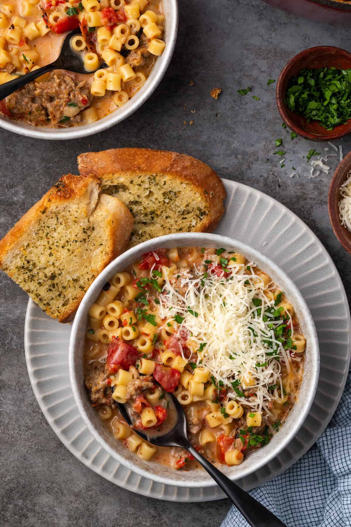 Overhead view of creamy Italian sausage soup in a bowl with a spoon, garnished with grated parmesan cheese, next to slices of garlic bread on a plate.