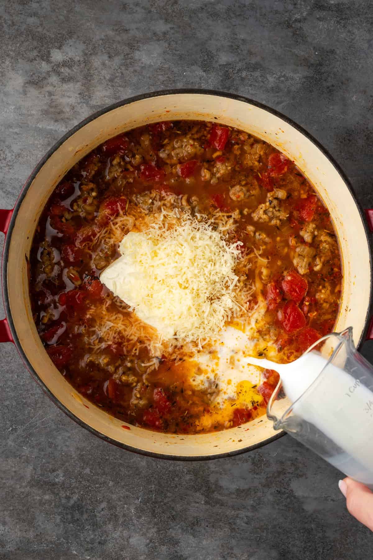 Milk being poured into a large pot of Italian sausage soup along with cream cheese and parmesan cheese.