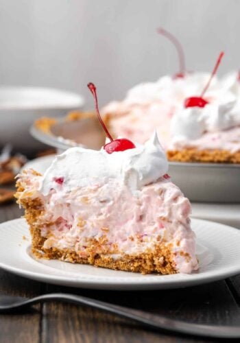 A slice of millionaire pie topped with whipped cream and a cherry on a plate, with the rest of the pie in the background.