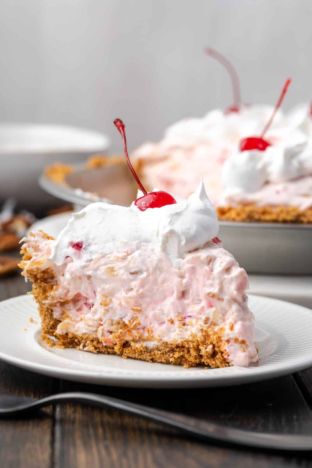 Millionaire pie is a decadent and fruity no-bake pie filled with sweet crushed pineapple, maraschino cherries, crispy pecans, and coconut. A slice of millionaire pie topped with whipped cream and a cherry on a plate, with the rest of the pie in the background.