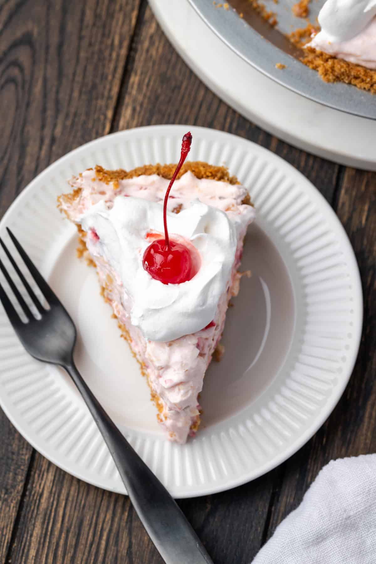 Millionaire pie is a decadent and fruity no-bake pie filled with sweet crushed pineapple, maraschino cherries, crispy pecans, and coconut. Overhead view of a slice of millionaire pie topped with whipped cream and a cherry on a plate.