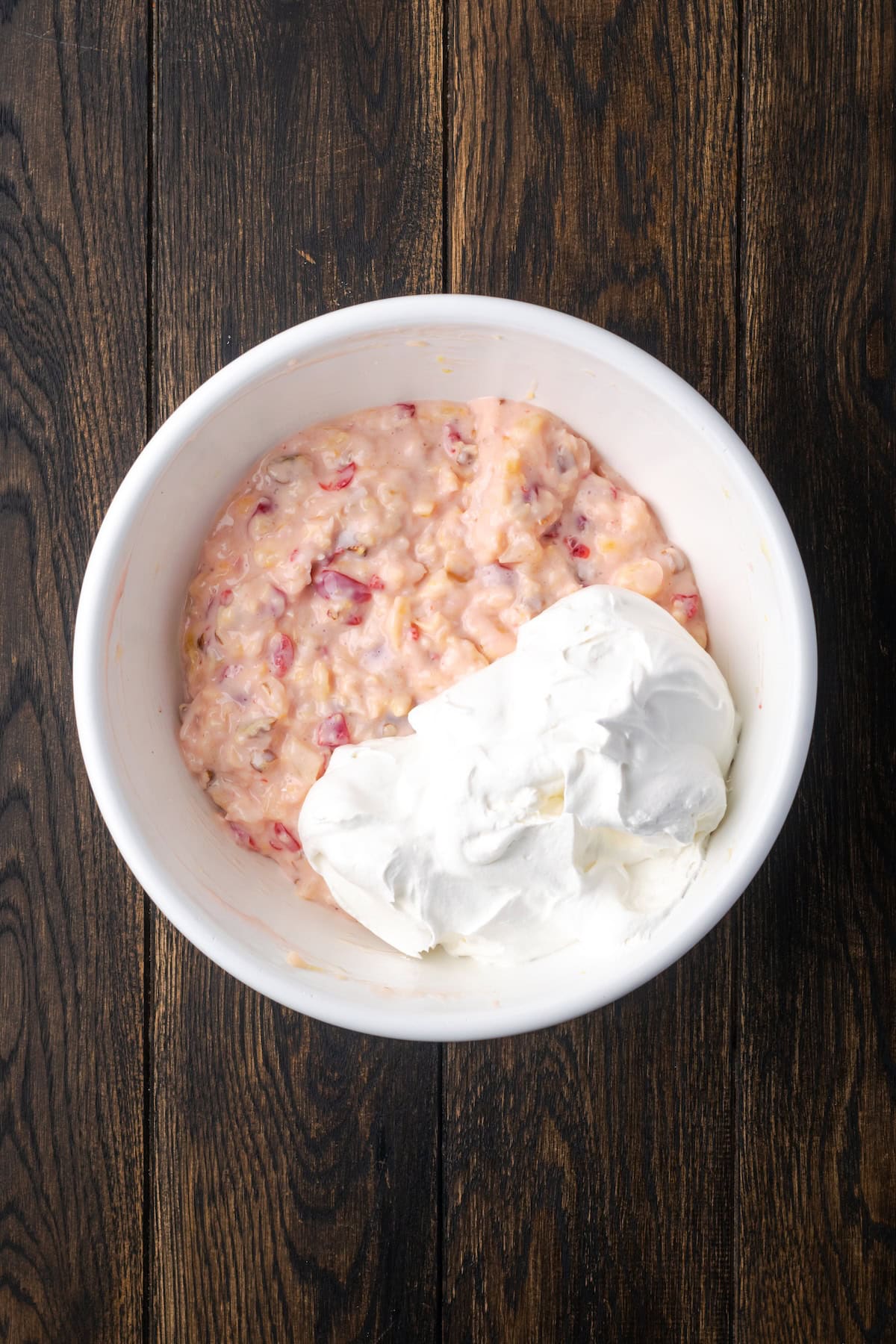 Millionaire pie is a decadent and fruity no-bake pie filled with sweet crushed pineapple, maraschino cherries, crispy pecans, and coconut. Whipped cream added to millionaire pie filling in a white bowl.