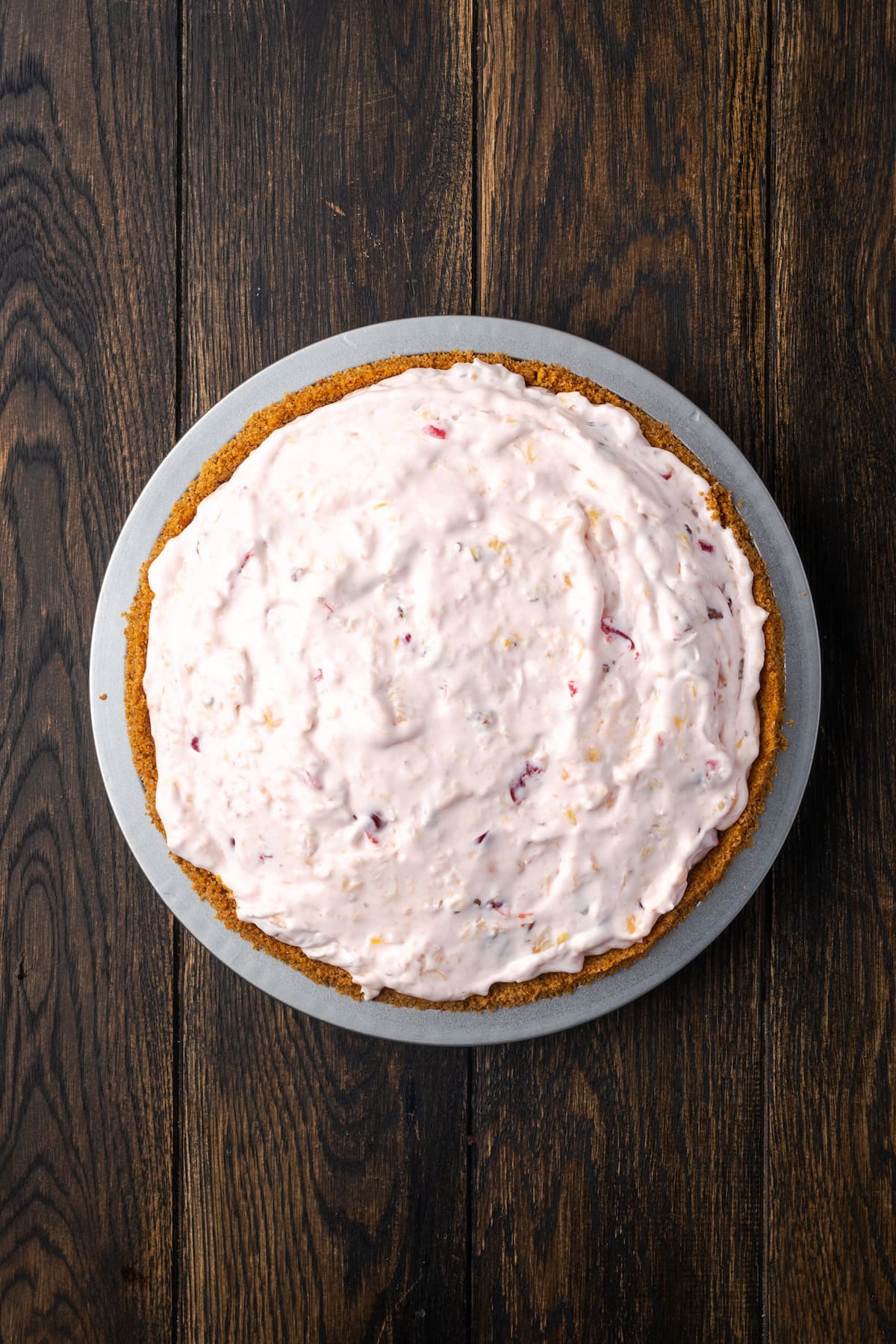 Millionaire pie is a decadent and fruity no-bake pie filled with sweet crushed pineapple, maraschino cherries, crispy pecans, and coconut. Overhead view of the assembled millionaire pie in a graham cracker crust.