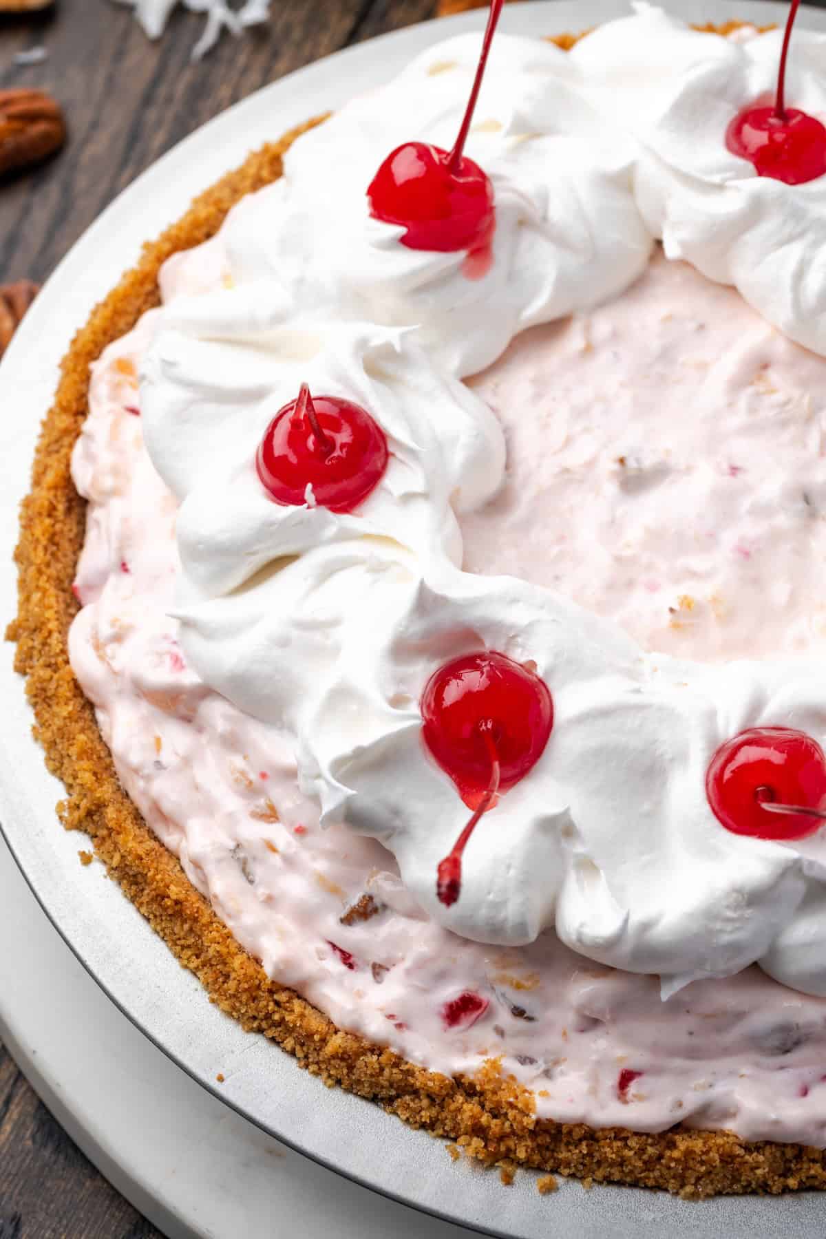 Millionaire pie is a decadent and fruity no-bake pie filled with sweet crushed pineapple, maraschino cherries, crispy pecans, and coconut. Close up of millionaire pie decorated with swirls of whipped cream and whole maraschino cherries.