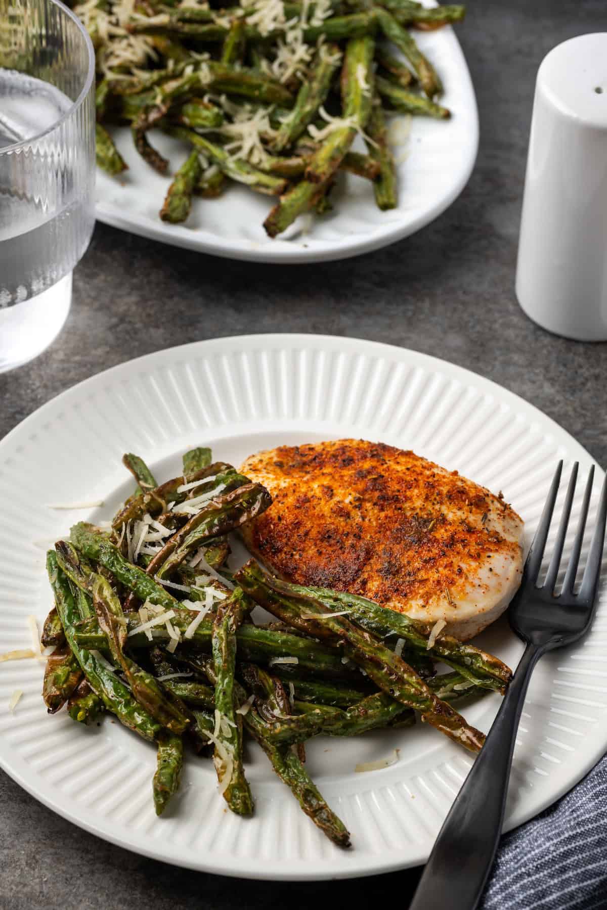 Air fryer green beans served beside a baked chicken breast on a plate next to a fork, with a platter of green beans in the background.