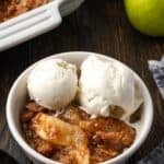 A serving of apple brown betty in a bowl topped with ice cream, with a baking dish in the background.