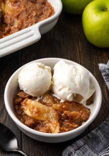 A serving of apple brown betty in a bowl topped with ice cream, with a baking dish in the background.