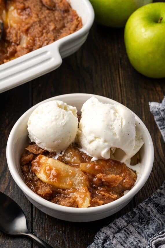 A serving of apple brown betty in a bowl topped with ice cream, with a baking dish in the background.