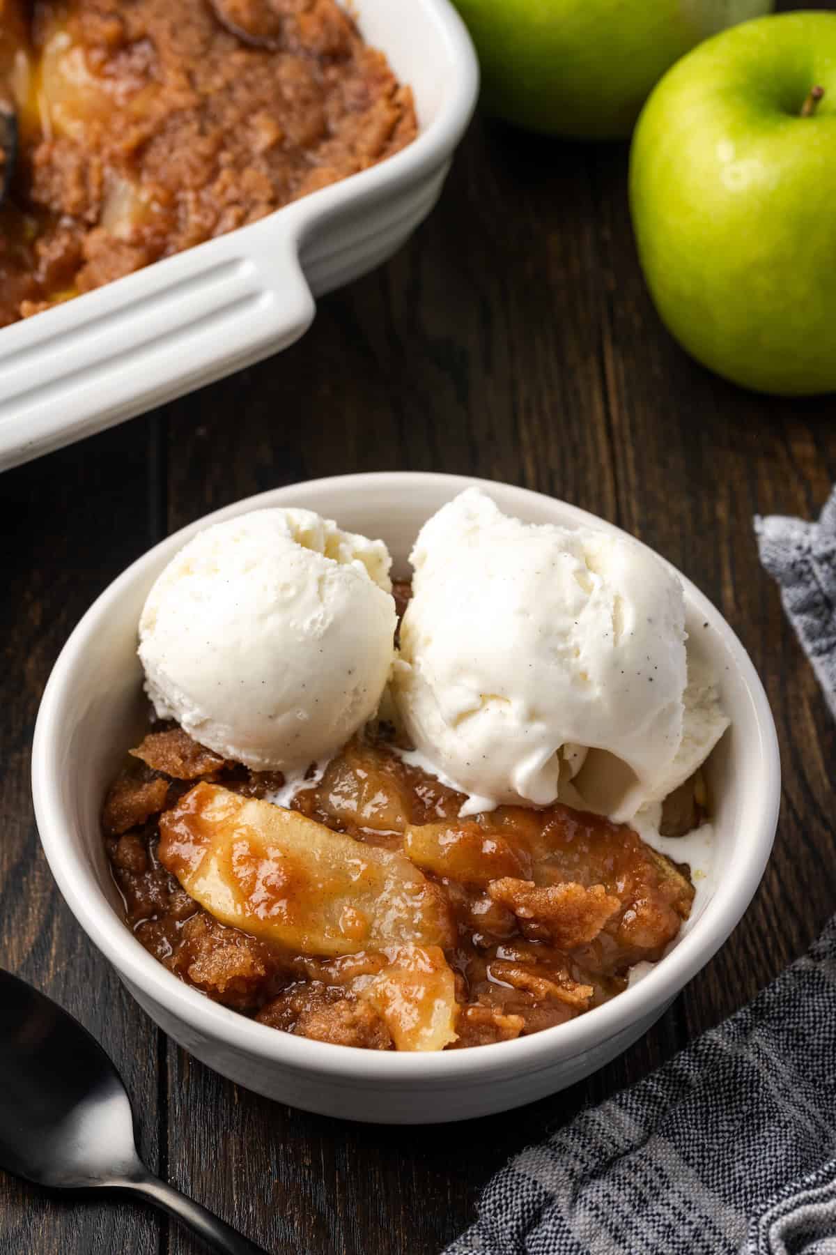 A serving of apple brown betty in a bowl topped with ice cream, with a baking dish in the background.