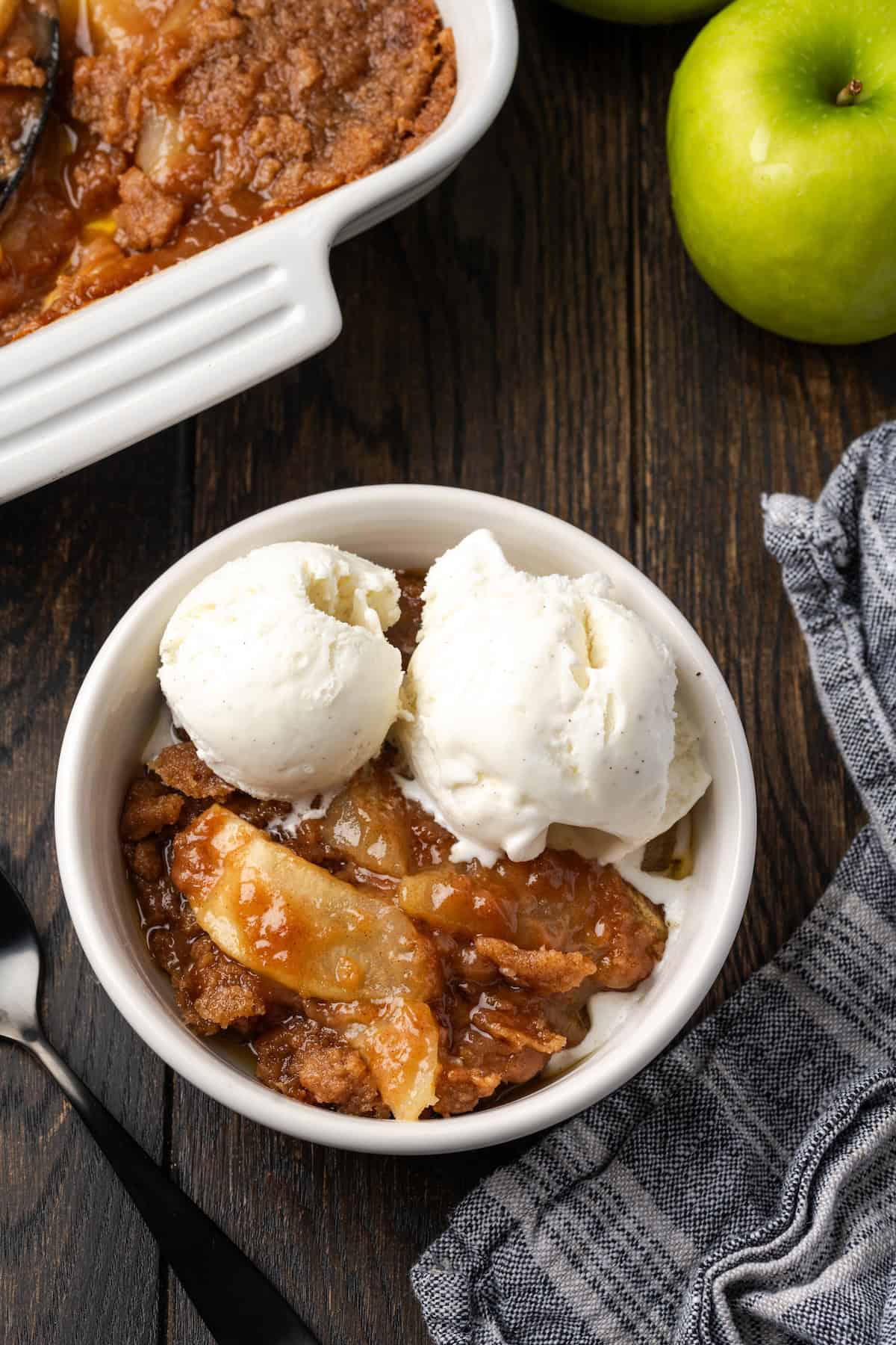 Overhead view of a serving of apple brown betty in a bowl topped with ice cream, next to a baking dish.