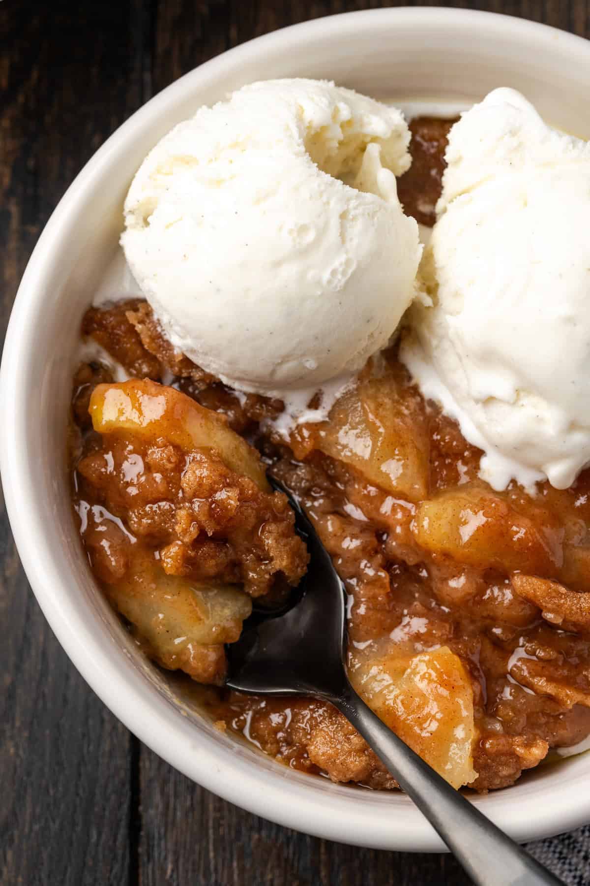 Overhead view of a serving of apple brown betty in a bowl topped with scoops of ice cream, with a spoon.