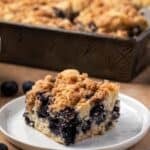 A slice of blueberry buckle on a plate with the rest of the cake in a baking pan in the background.