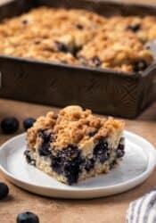 A slice of blueberry buckle on a plate with the rest of the cake in a baking pan in the background.