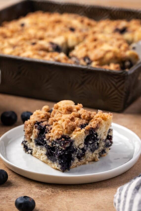 A slice of blueberry buckle on a plate with the rest of the cake in a baking pan in the background.
