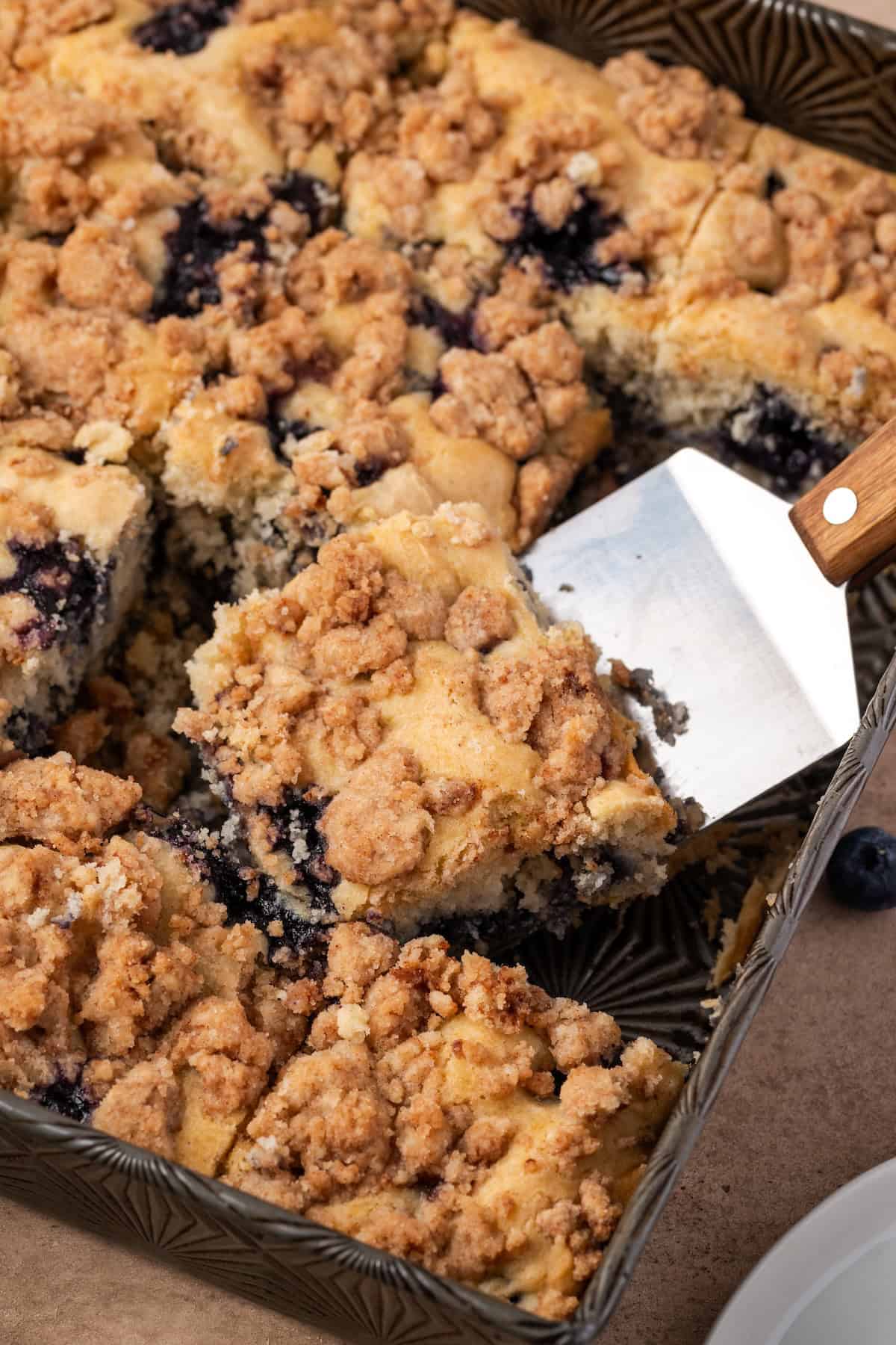 A cake server lifting a slice of blueberry buckle from a baking dish.