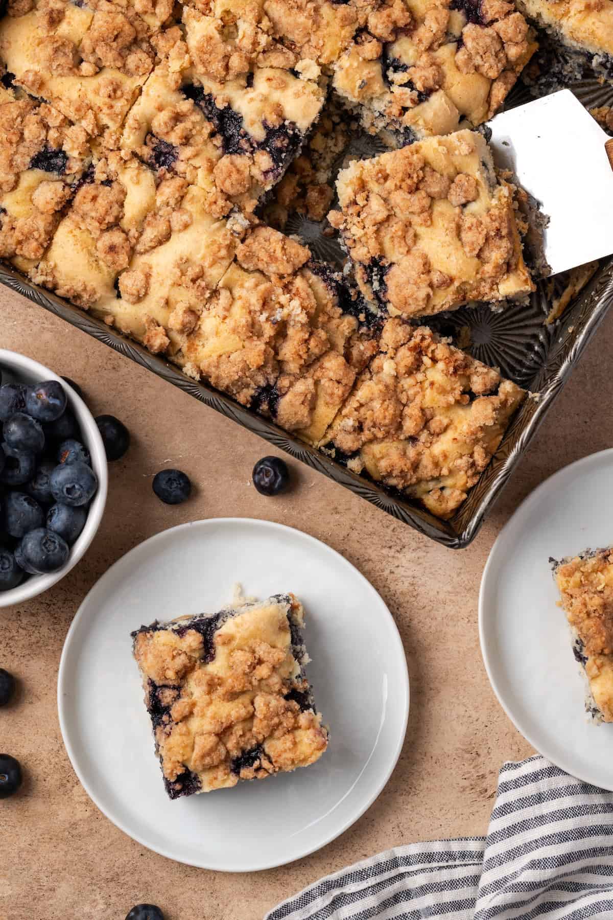 Overhead view of a slice of blueberry buckle on a plate next to the rest of the cake in a baking pan.