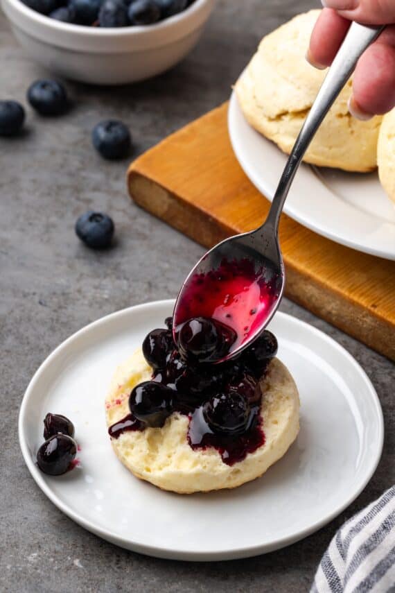 A spoon adding blueberry sauce to one half of a shortcake biscuit on a plate.
