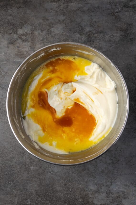 This red velvet cheesecake has four decadent layers of creamy cheesecake and soft red velvet cake smothered with cream cheese frosting. Eggs and vanilla added to cheesecake batter in a bowl.