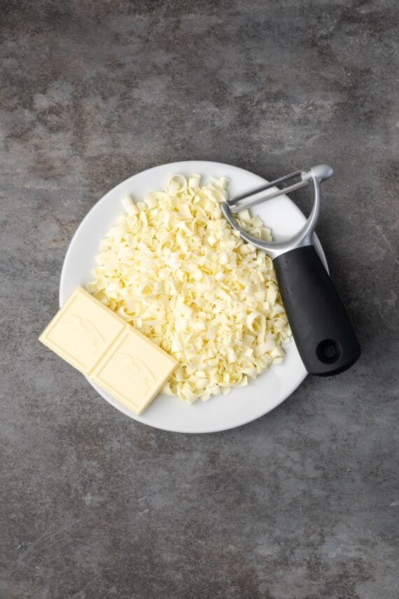 This red velvet cheesecake has four decadent layers of creamy cheesecake and soft red velvet cake smothered with cream cheese frosting. Shaved white chocolate curls on a plate next to chocolate squares and a peeler.