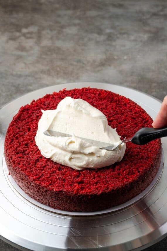 This red velvet cheesecake has four decadent layers of creamy cheesecake and soft red velvet cake smothered with cream cheese frosting. An offset spatula spreading frosting over the first red velvet cake layer.