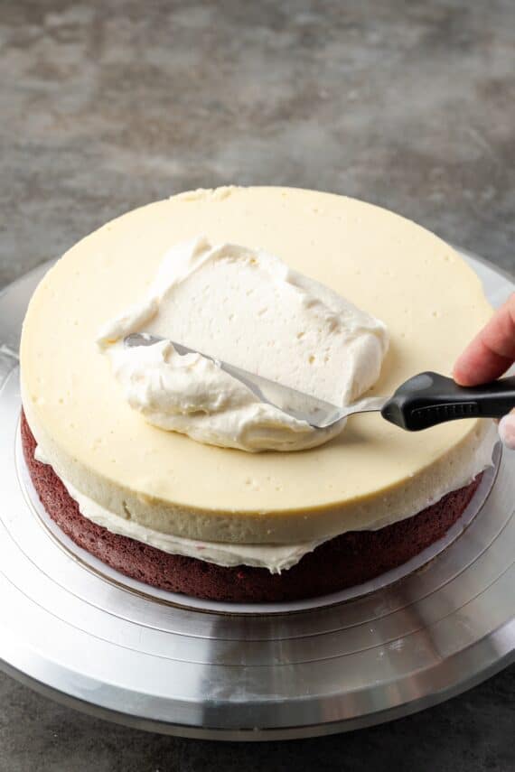 This red velvet cheesecake has four decadent layers of creamy cheesecake and soft red velvet cake smothered with cream cheese frosting. An offset spatula spreading frosting over the cheesecake layer.