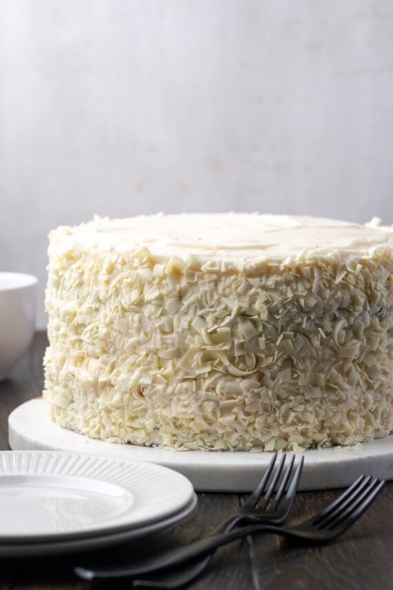 This red velvet cheesecake has four decadent layers of creamy cheesecake and soft red velvet cake smothered with cream cheese frosting. Frosted red velvet cheesecake decorated with white chocolate curls on a plate.