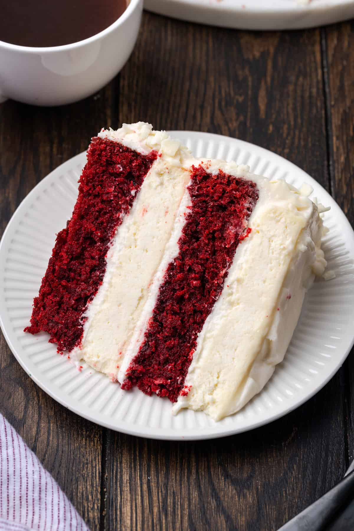 This red velvet cheesecake has four decadent layers of creamy cheesecake and soft red velvet cake smothered with cream cheese frosting. A slice of red velvet cheesecake on a plate with a cup of coffee in the background.
