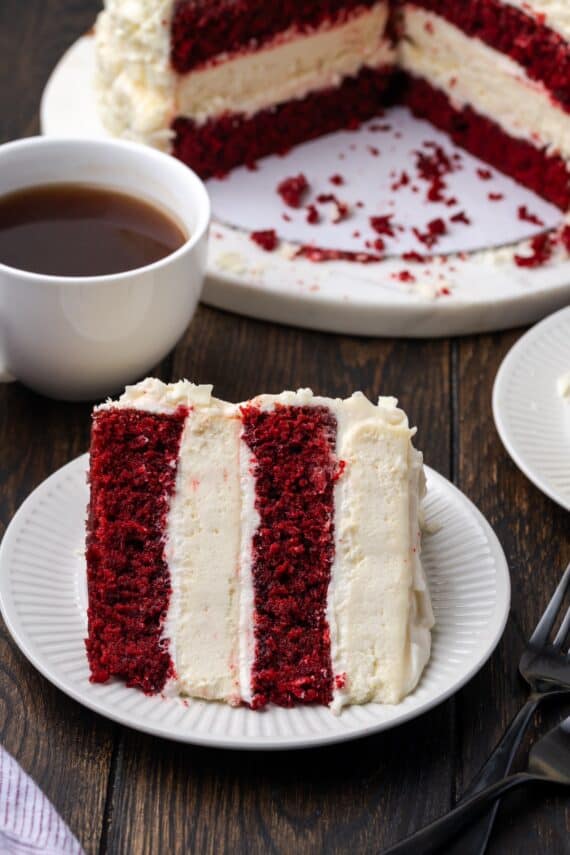 A slice of red velvet cheesecake on a plate with a cup of coffee and the rest of the cheesecake in the background.