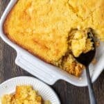 Overhead view of spoon bread in a baking dish with a spoon scooping from the corner, next to a serving on a plate.