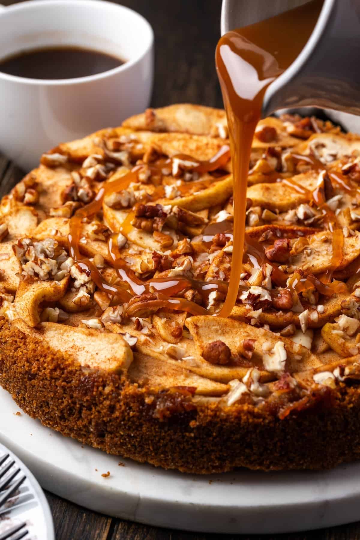This homemade apple cheesecake recipe has a buttery graham cracker crust layered with creamy cheesecake and a gooey caramel apple topping. Caramel sauce being poured over an apple cheesecake garnished with pecans.