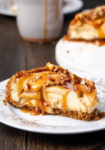 A slice of apple cheesecake covered with caramel sauce on a plate with the rest of the cheesecake in the background.