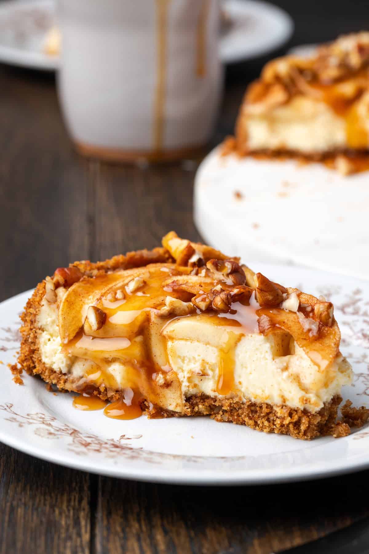 This homemade apple cheesecake recipe has a buttery graham cracker crust layered with creamy cheesecake and a gooey caramel apple topping. A slice of apple cheesecake covered with caramel sauce on a plate with the rest of the cheesecake in the background.
