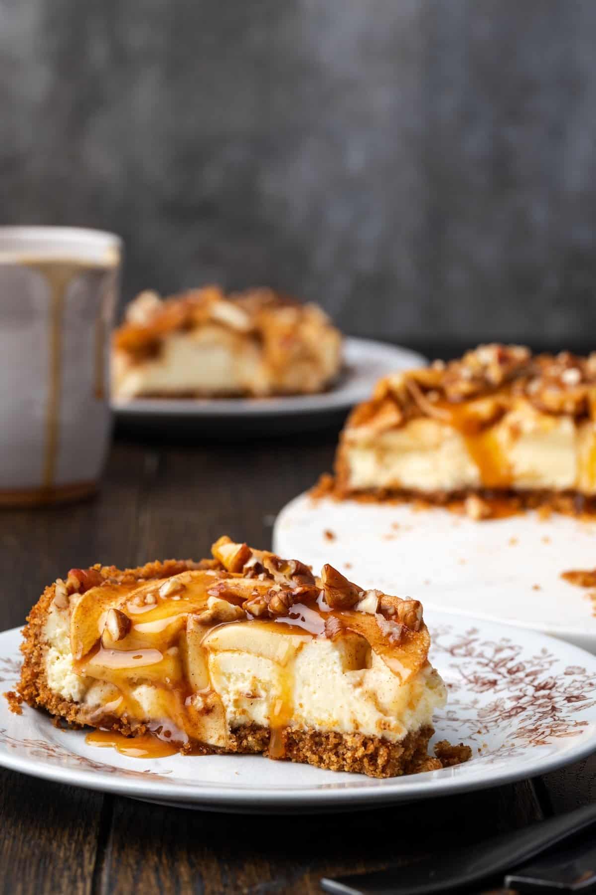 This homemade apple cheesecake recipe has a buttery graham cracker crust layered with creamy cheesecake and a gooey caramel apple topping. A slice of apple cheesecake covered with caramel sauce on a plate with more cheesecake slices in the background.