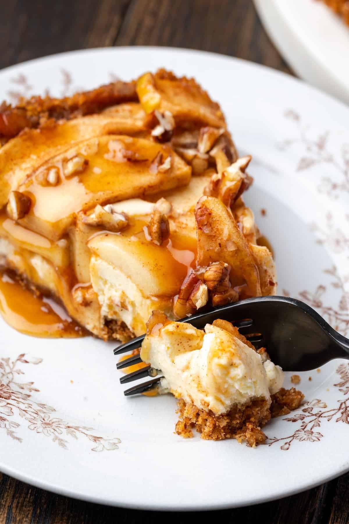 This homemade apple cheesecake recipe has a buttery graham cracker crust layered with creamy cheesecake and a gooey caramel apple topping. A fork cutting into a slice of apple cheesecake on a plate.