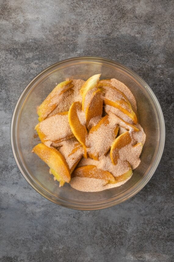 This homemade apple cheesecake recipe has a buttery graham cracker crust layered with creamy cheesecake and a gooey caramel apple topping. Apple slices tossed with cinnamon sugar in a bowl.