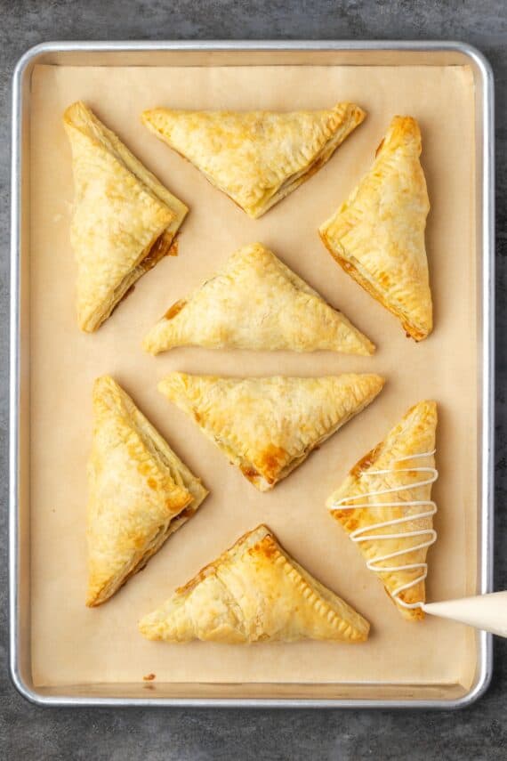 Crispy, buttery store-bought puff pastry and quick, homemade apple filling make these easy apple turnovers the perfect fall weekend treat! Overhead view of a piping bag piping icing over an apple turnover, next to more turnovers on a baking sheet.