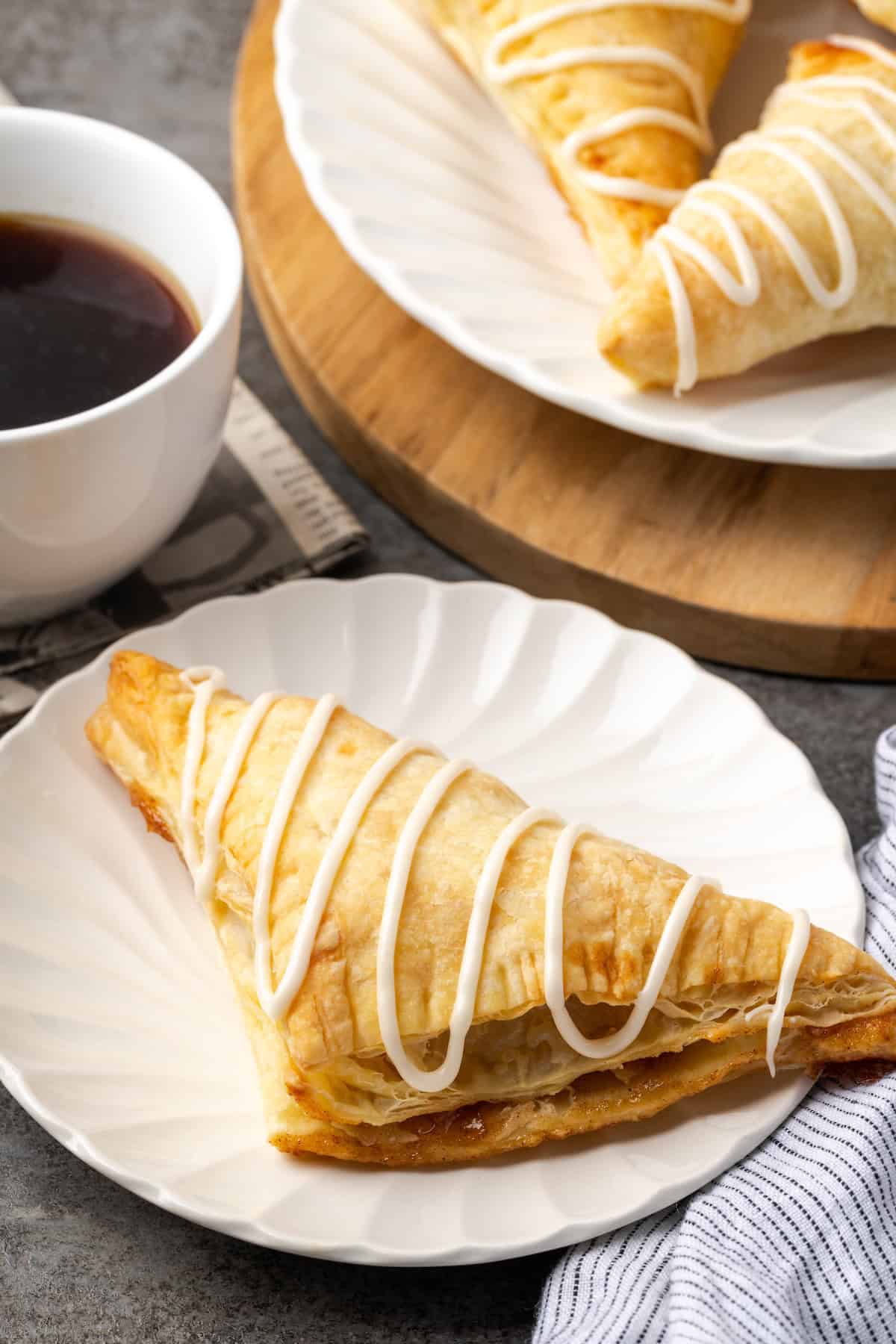 Crispy, buttery store-bought puff pastry and quick, homemade apple filling make these easy apple turnovers the perfect fall weekend treat! An apple turnover drizzled with icing on a plate next to a cup of coffee and another plate of turnovers.