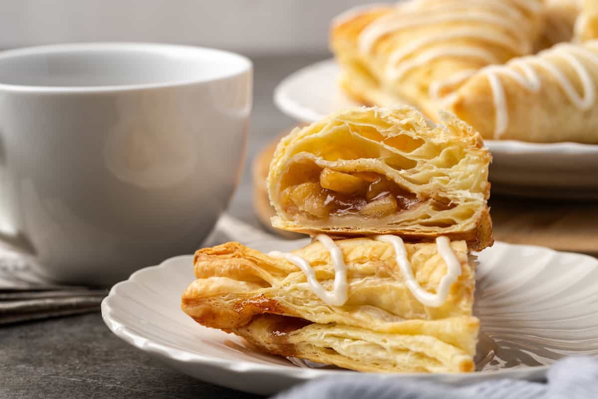 Crispy, buttery store-bought puff pastry and quick, homemade apple filling make these easy apple turnovers the perfect fall weekend treat! Two halves of an apple turnover stacked on a plate with a coffee cup and more turnovers in the background.