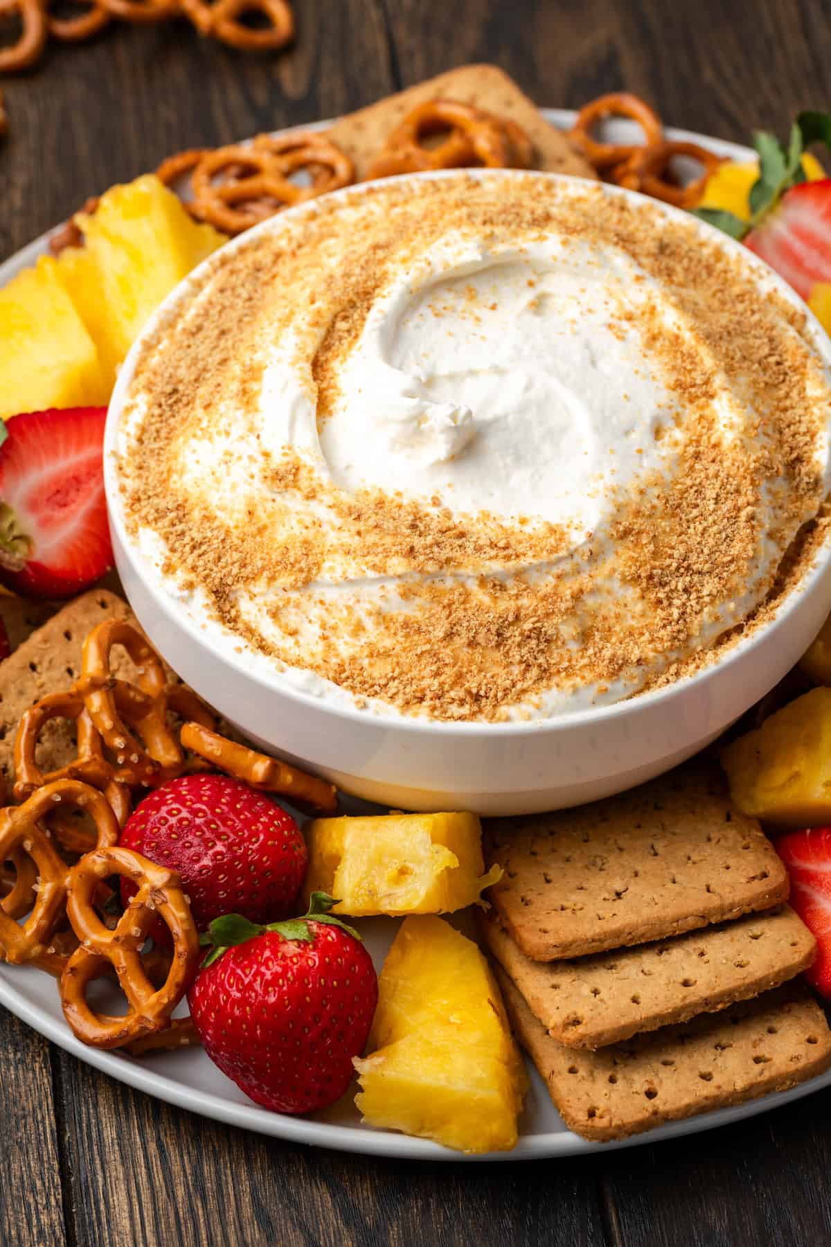A bowl of cheesecake dip garnished with graham cracker crumbs in the center of a large platter of fresh fruit, pretzels, and graham crackers.